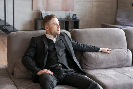 A man in a formal suit is sitting on a gray sofa in a modern office, relaxed, with a focused look, expressing calmness and confidence.の写真素材