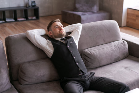 man in business clothes is sitting relaxed on the couch with his hands behind his head, with thoughtful expression on his face. modern interior and soft lighting create an atmosphere calm and reflectionの写真素材