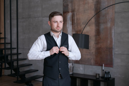 A man in an elegant suit with a vest is buttoning up, standing in a modern interior with a staircase...