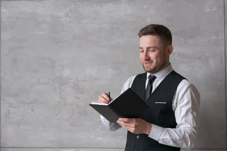 man in business clothes stands against wall holding notebook and pen in his hands concentrated expression on his face. neutral background, soft lighting emphasize atmosphere professionalism.の写真素材