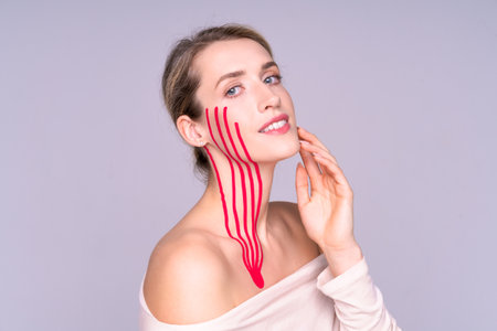 young smiling woman with red tapes on her face and neck. She gently touches her face with her hand, aesthetic cosmetology, skin care, rejuvenation, beauty without surgery, self-care.の写真素材