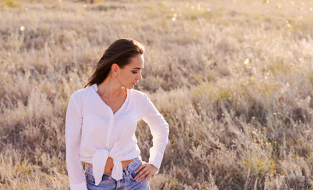 Woman is standing in a field wearing a white shirt and blue jeans. She is looking to her rightの写真素材