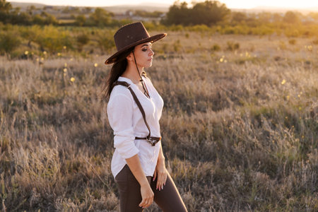 Woman wearing a brown hat and a white shirt is standing in a field. She is wearing a brown belt and a brown vestの写真素材