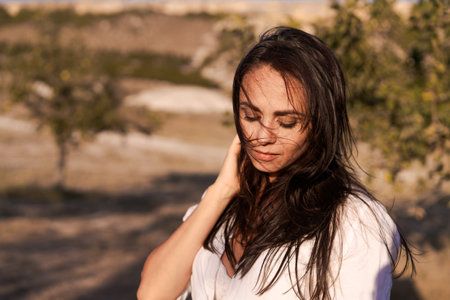 Woman with long hair is standing in the sun. She is wearing a white shirt and has her hair blowing...