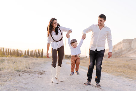 Family of three, a man, a woman, and a child, are walking on a dirt road. The man is holding the child, who is hanging from his arm. The woman is walking behind them, and they are all smilingの写真素材