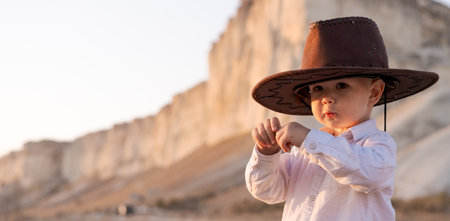 Baby wearing a cowboy hat. The baby is standing in front of a large rock. The baby is wearing a white shirtの写真素材
