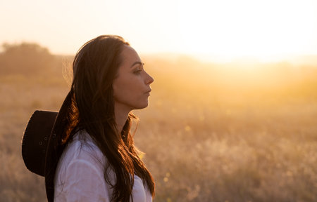 Woman is standing in a field with a hat on her head. She is looking at the sunの写真素材