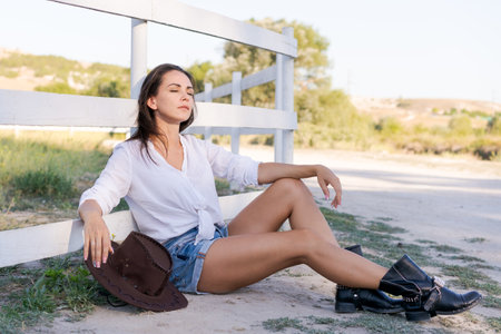 Woman is sitting on the ground wearing a cowboy hat and shorts. She is wearing boots and a white shirtの写真素材