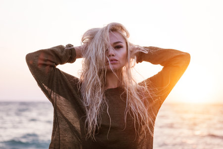 Blonde woman with long hair is standing on the beach. She is wearing a green shirt and has her arms...