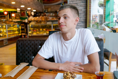 Young man is sitting at a table in a restaurant. He is wearing a white shirt and smilingの写真素材