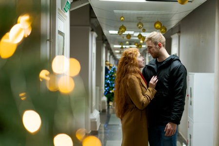 Man and a woman are standing in a hallway. The man is wearing a black jacket. The woman is wearing a...