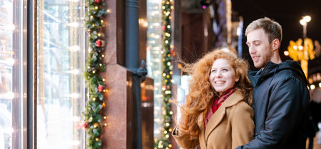 Man and a woman are standing in front of a window with Christmas decorations. The woman has red hair and is wearing a scarfの写真素材