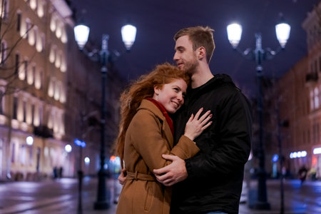 Man and a woman are hugging each other on a street. The man is wearing a black jacket and the woman is wearing a brown coatの写真素材