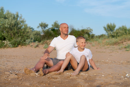Man and a boy are sitting on the beach. The boy is wearing a white shirtの写真素材