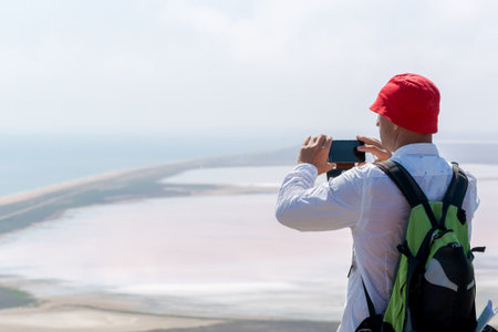 Man wearing a red hat is taking a picture of the ocean. He is holding a cell phone in his handの写真素材