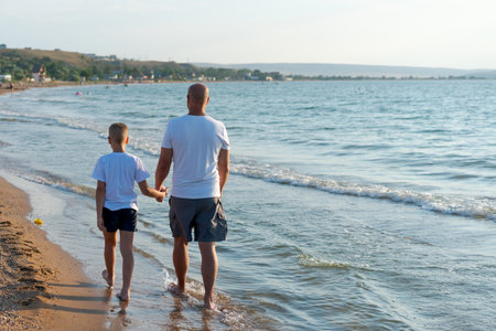 Man and a boy are walking on the beach. The man is holding the boy's handの写真素材