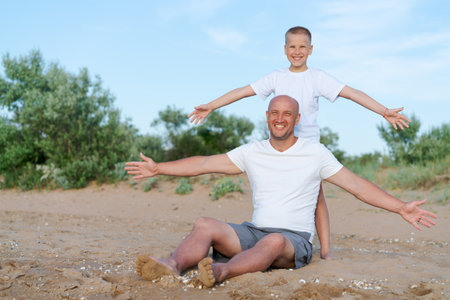 Man and a boy are sitting on the sand. The man is wearing a white shirt and shortsの写真素材