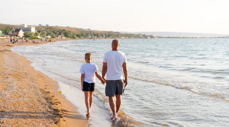 Man and a boy are walking on the beach. The boy is wearing a white shirt. The man is holding the boy's handの写真素材