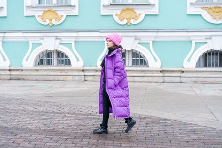 Woman wearing a pink coat and a pink hat. She is walking on a brick sidewalk. The brick sidewalk is next to a building with a white facadeの写真素材