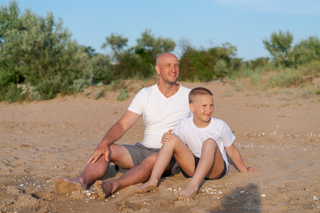 Man and a boy are sitting on the beach. The man is wearing a white shirt and shortsの写真素材