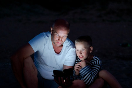 Man and a boy are sitting on the ground looking at a cell phone. The boy is wearing a striped shirtの写真素材