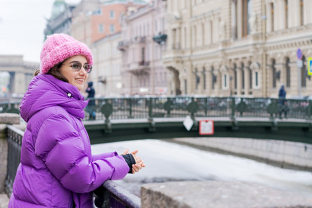 A woman in a purple coat and pink hat stands on a bridge. She is smiling and looking out over the waterの写真素材