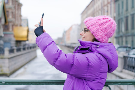 A woman wearing a pink hat and a purple coat is taking a picture of herself with her cell phoneの写真素材