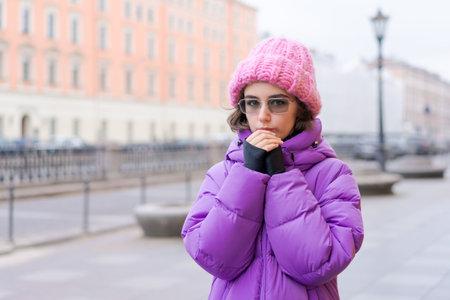 A girl wearing a pink hat and a purple coat is standing on a sidewalk. She is shivering and she is coldの写真素材