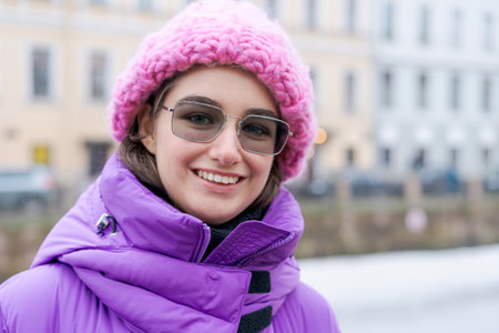 A woman wearing a pink hat and glasses is smiling. She is standing in front of a building