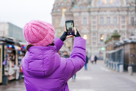 A woman wearing a pink hat is taking a picture of a building with a cell phone. Concept of adventure and exploration, as the woman is capturing a moment in a new placeの写真素材