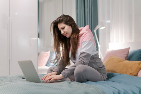 A woman is sitting on a bed with a laptop in front of her. She is typing on the laptop and she is focused on her work. The room is well-lit and has a cozy atmosphere, with a few pillowsの写真素材
