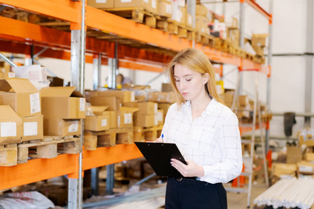 Young focused woman supervisor in a white shirt writing on a clipboard in a large logistics warehouse. Background with orange metal racks and cardboard boxes. Business and distribution concept.の写真素材