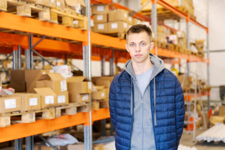 A focused young man standing in a large distribution warehouse with orange racks, wooden pallets, and stock boxes. Concept of logistics, storage, and e-commerce inventory.の写真素材