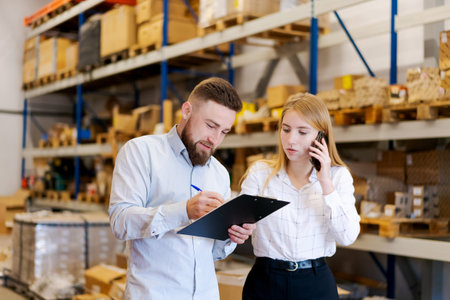 Professional male supervisor writing on a clipboard while his female colleague manages a phone call in a distribution center. Concept of documentation, teamwork, and supply chain management.の写真素材