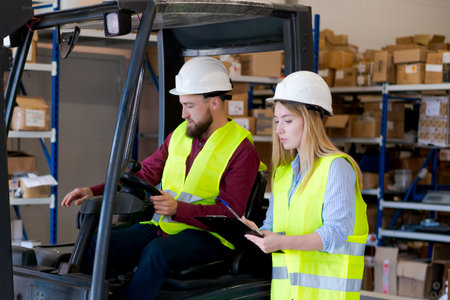 Logistics team in reflective vests and hard hats. Female supervisor with clipboard coordinating stock movement with a male forklift driver in a distribution center.の写真素材
