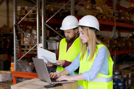 Male worker reviewing document while female colleague processes information on laptop. Business intelligence and performance metrics in logistics managementの写真素材