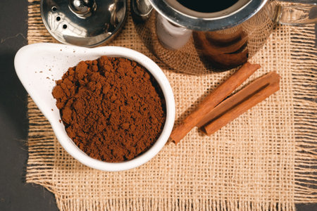 Cocoa powder in a bowl with cinnamon and coffee maker on sackcloth background