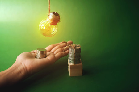 Hand holding a light bulb and stack of coins on green background with copy spaceの写真素材