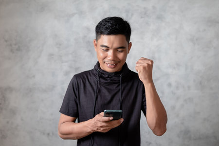 Confident excited young Asian man looking at smartphone isolated on gray background. Winner conceptの写真素材