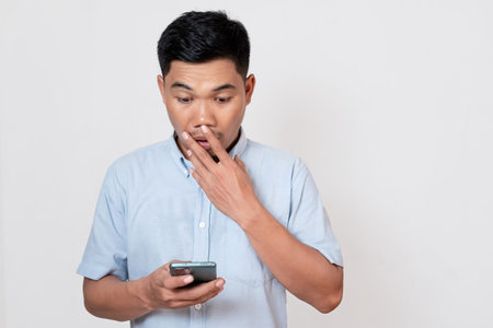 Confident Asian young man holding smartphone covering mouth with hand, shocked and scared of mistake. surprised expressionの写真素材