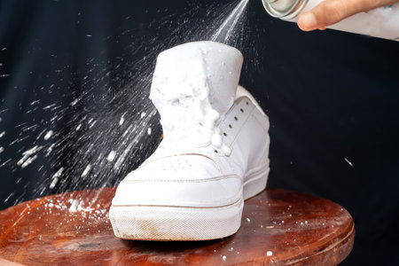 A woman sprays cleansing foam onto white leather shoes. Care for leather footwear.
