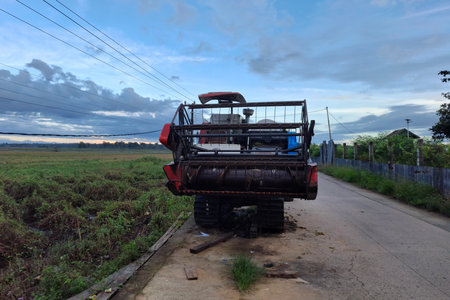 RedAndWhiteAgriculturalVehicleParkedOnDirtRoadNearFieldAtSunsetWithCloudySkyの写真素材