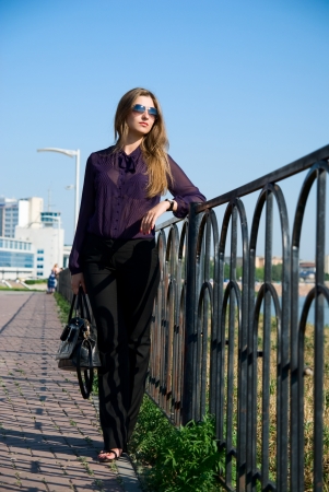 The beautiful girl with long hair in dark glasses against embankment.Holds a ladies' handbagの写真素材