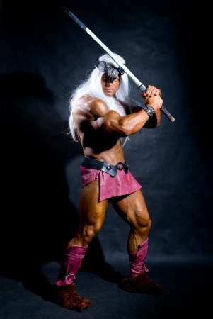 Muscular man in an image of a barbarian with a raised sword. On a dark background in full lengthの写真素材