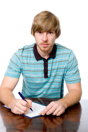 A young man sits at a desk with a pen in hand. Looking at the camera isolated on a white backgroundの写真素材