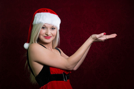 Santa girl in Christmas cap gestures palm up, isolated on redの写真素材