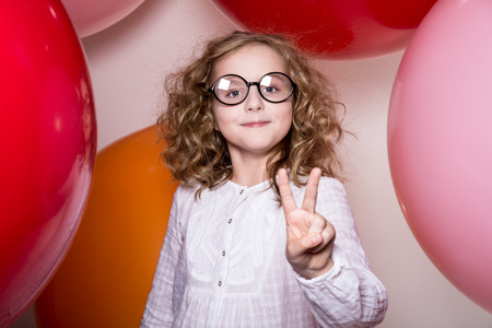 Young girl in glasses showing two fingers victory against the background of large colorful balloons.の写真素材