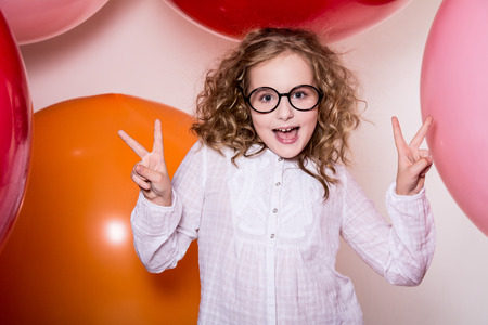 Young girl in glasses showing two fingers victory against the background of large colorful balloonsの写真素材