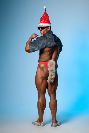 Strong athletic man in a santa hat and dark glasses back. Bodybuilder on a blue background. Christmas fancy dressの写真素材