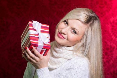 A girl in a white sweater and striped gift with white bow looking at the camera. Close-up portrait of a beautiful young woman on a red background. Holiday Saleの写真素材